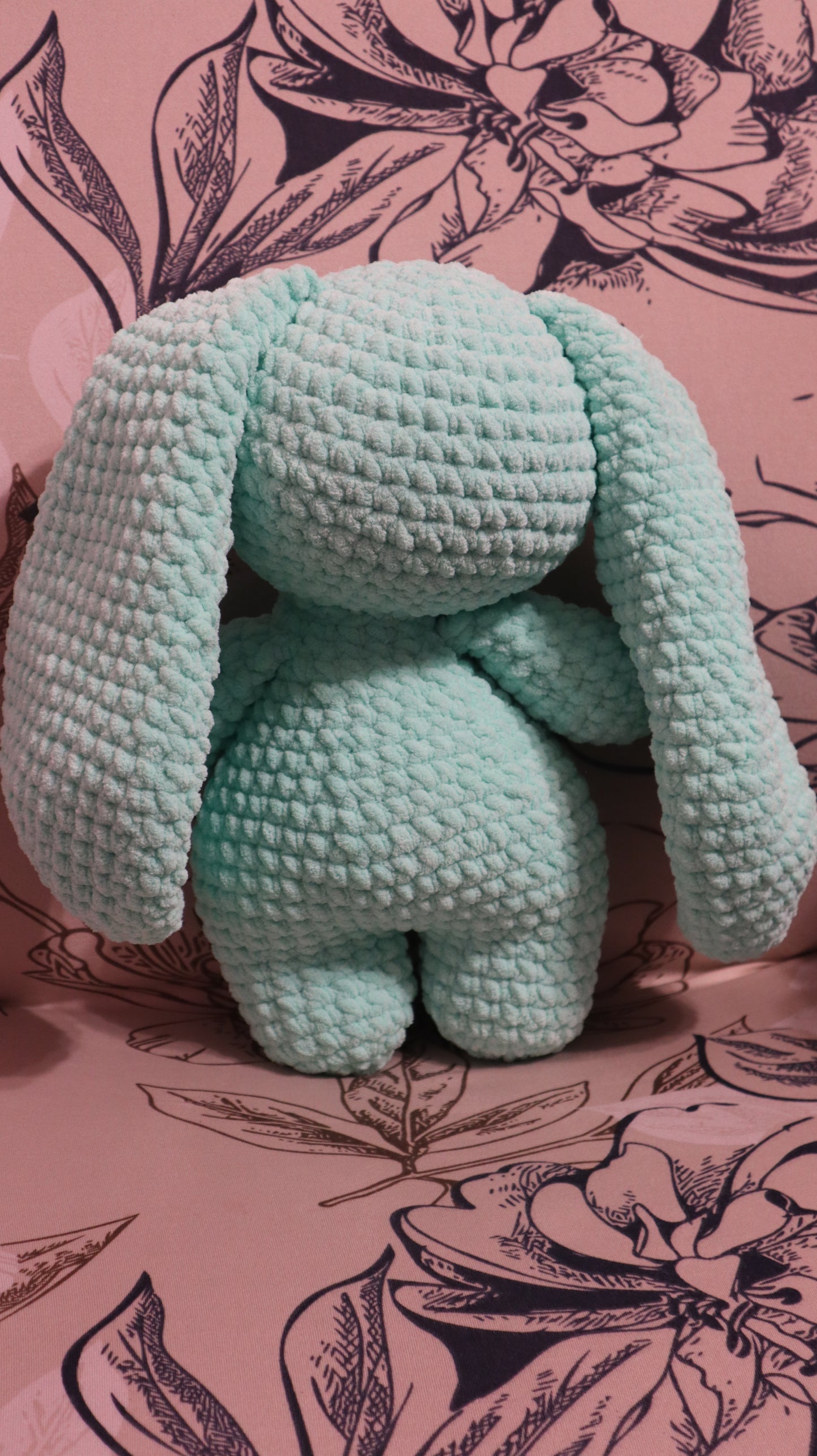 Back view of a crocheted seafoam green bunny plushie on a floral fabric background