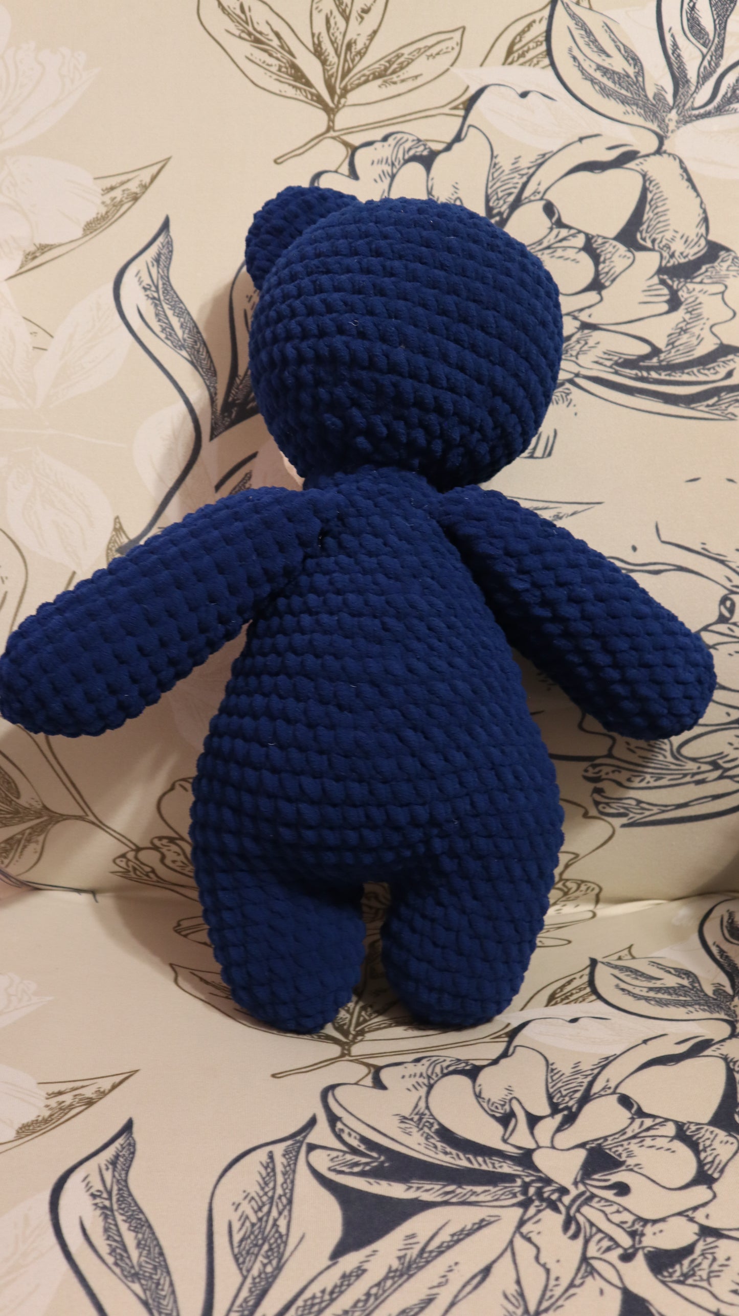 Back view of a dark Blue crocheted bear on a floral-patterned fabric background