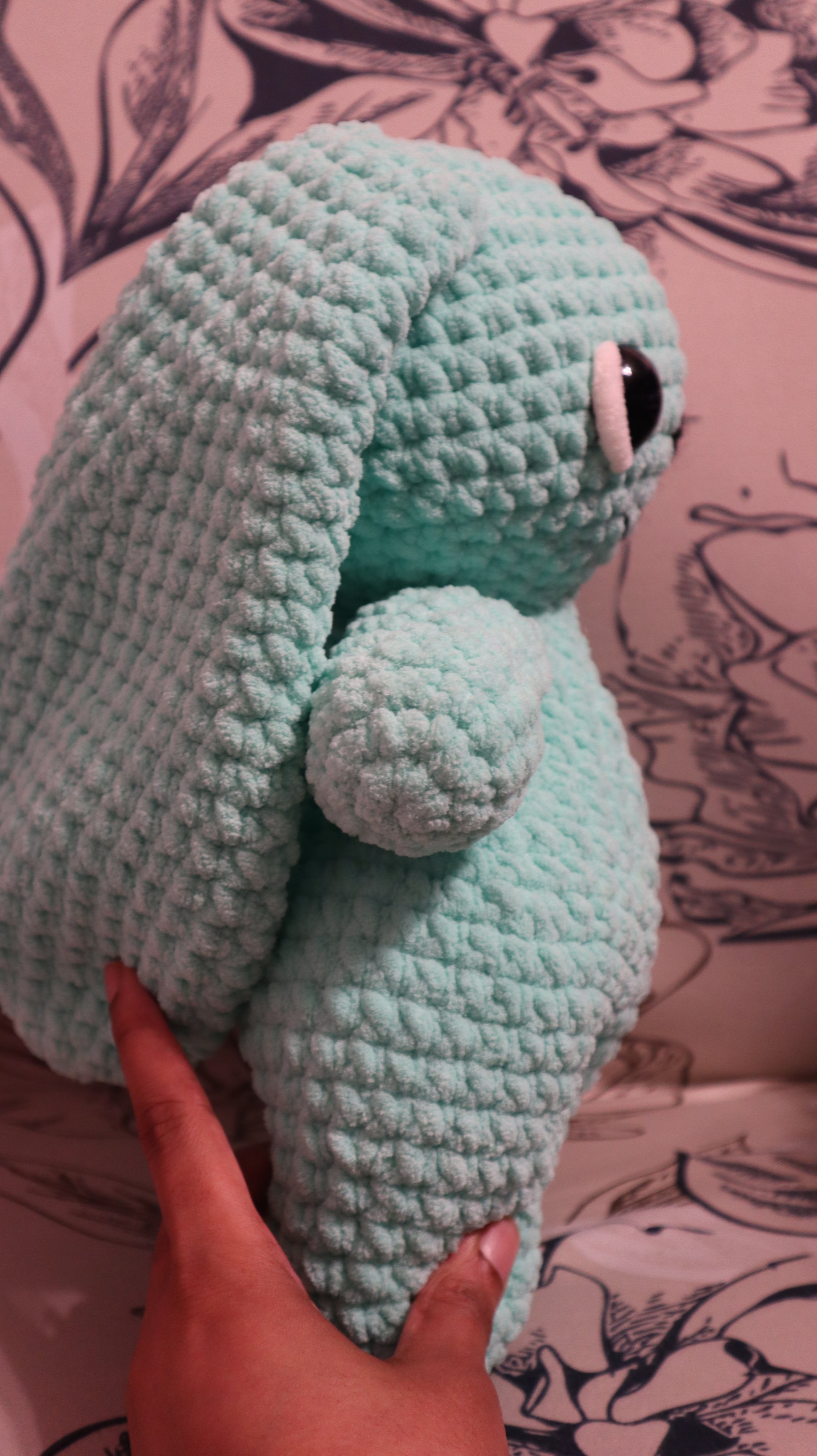 Side view of a crocheted seafoam green bunny plushie  on a floral fabric background