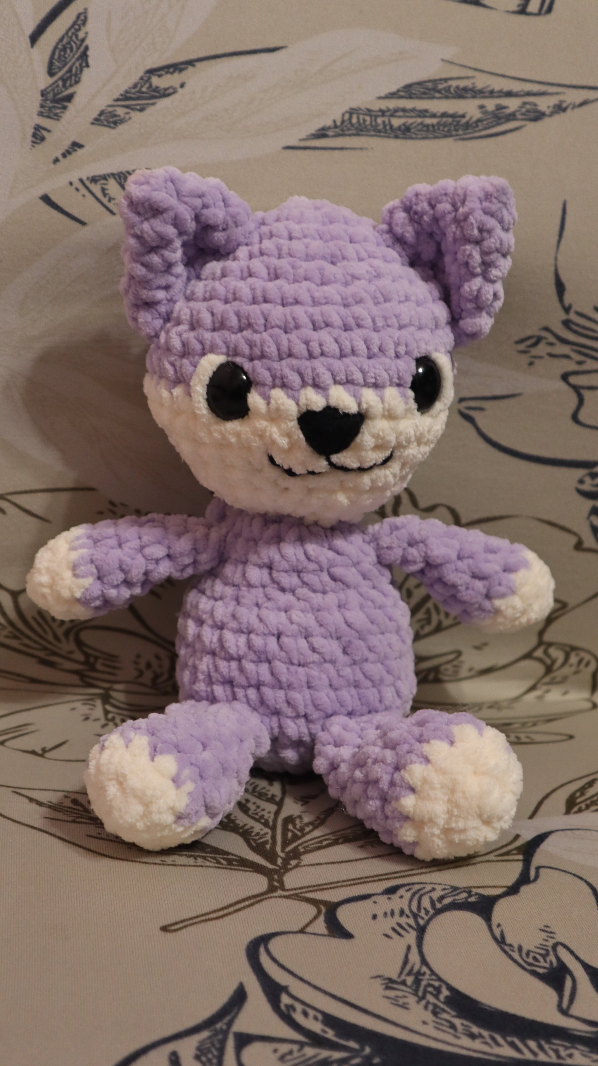 Crocheted purple and white kitten plushie on a floral fabric background