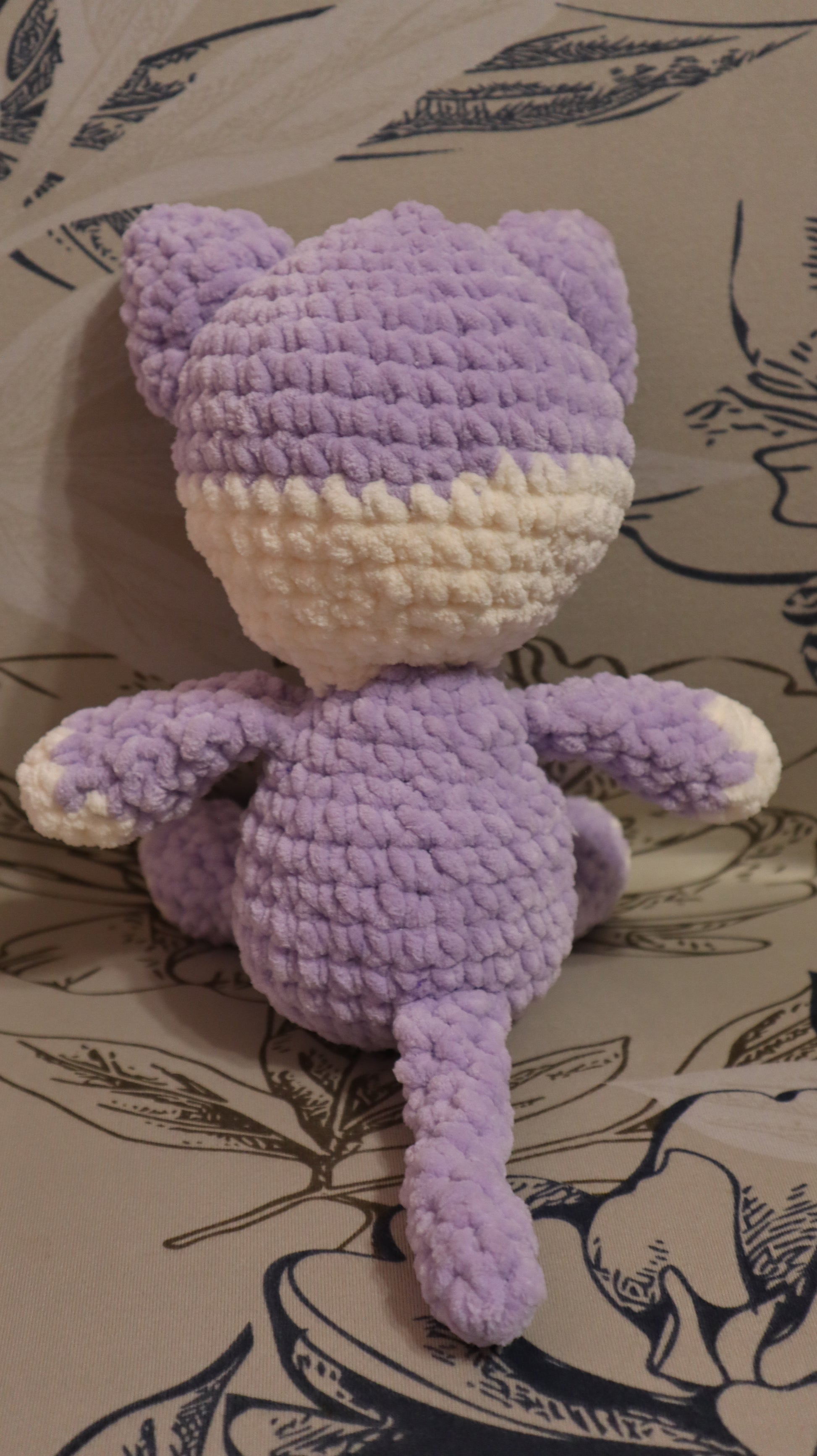 Back view of a crocheted purple and white kitten plushie on a floral fabric background