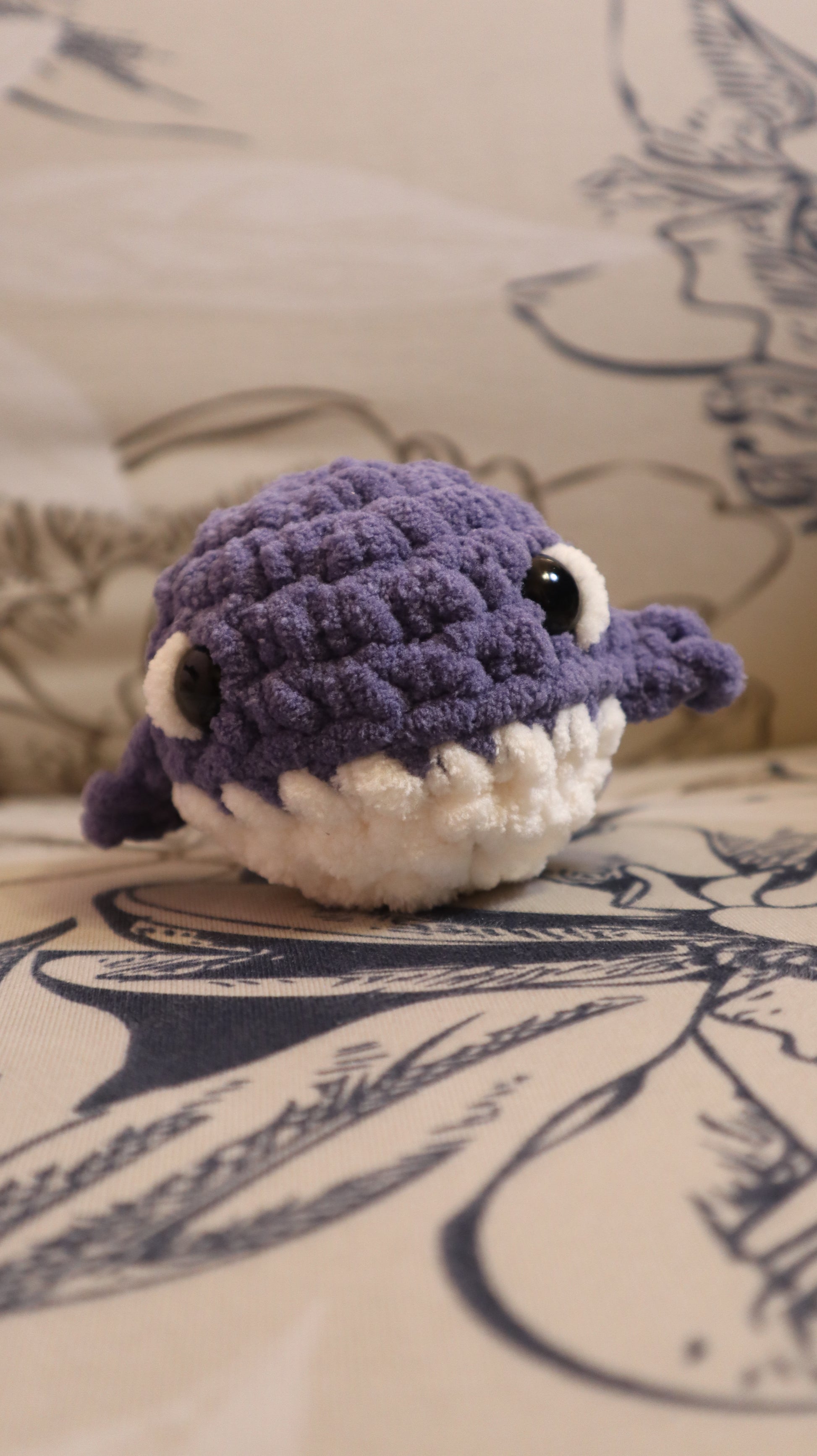 Dark Purple and white crocheted whale plushie on a floral pattern fabric background