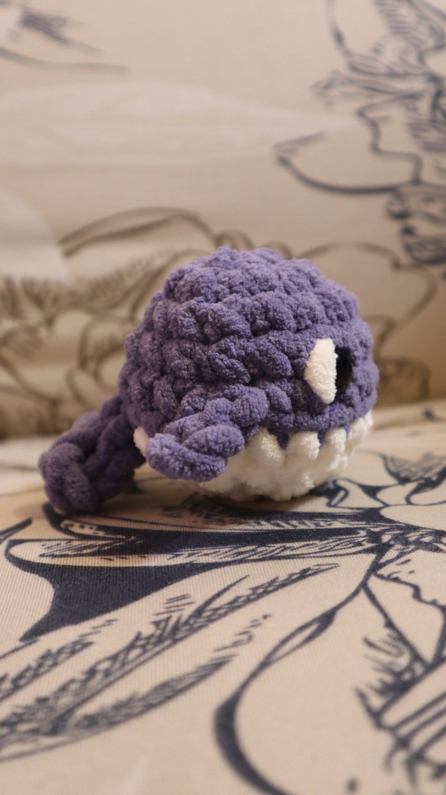 Side view of a dark purple and white crocheted whale plushie on a floral pattern fabric background
